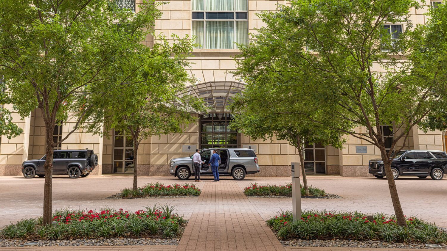 Two people at the entrance of a luxury hotel with glass canopy, flanked by trees, flower beds, and parked SUVs.