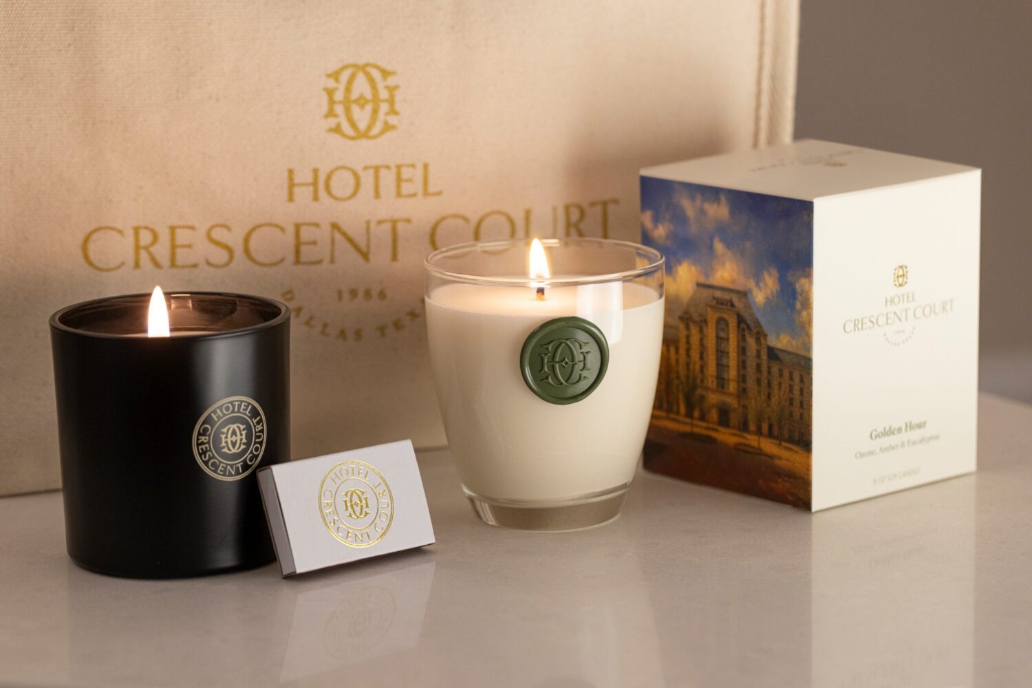 Two lit Hotel Crescent Court branded candles, matchbox, and gift box displayed against a beige branded fabric backdrop.