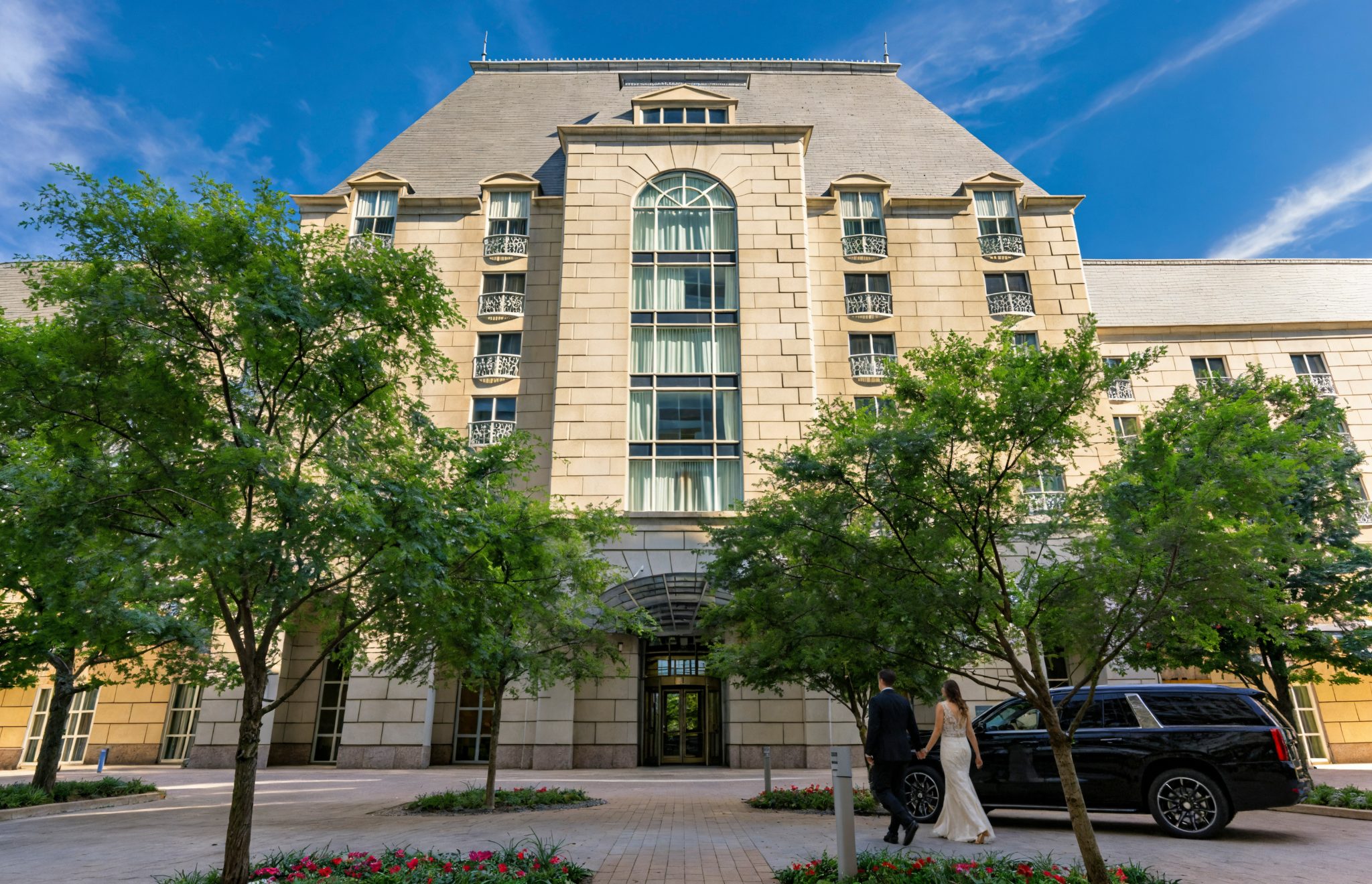 Bride and groom walking toward elegant stone luxury hotel entrance with arched windows and iron balconies in Dallas.
